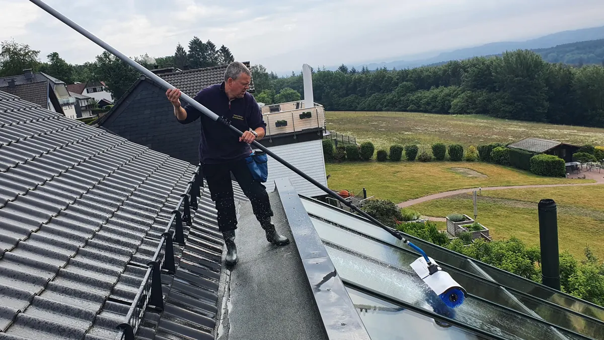 Mitarbeiter reinigt mit einer Teleskopstange die Dachverglasung eines Hauses, dahinter die Hügel des Sauerlands